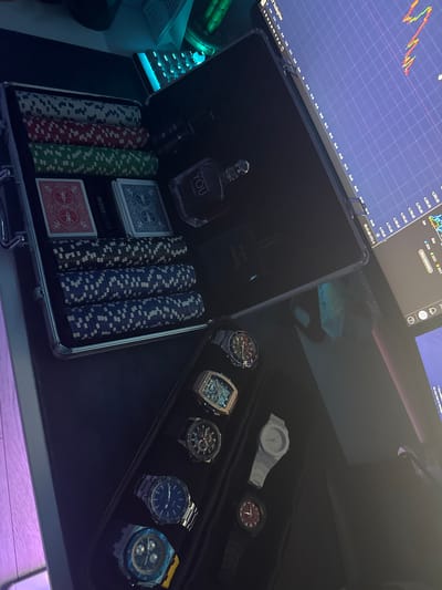 The image depicts a close-up, high-angle shot of a dark-colored open briefcase on a black surface. The briefcase contains several items, including stacks of poker chips in red, green, and blue, two decks of playing cards, a bottle of "YOU" perfume, and a bottle of "BLEU DE CHANEL" cologne. The briefcase is placed next to a computer monitor displaying a financial trading chart. Below the briefcase, a black watch organizer is visible, containing five wristwatches of various designs and colors. The background is dark, with hints of a keyboard illuminated with blue light and a glimpse of what appears to be a laptop or another screen. The overall scene suggests a personal collection or organized storage of luxury items, possibly related to gaming and personal grooming, set against a backdrop of financial monitoring. There is no discernible indication of the location in Busmantsi, Bulgaria.