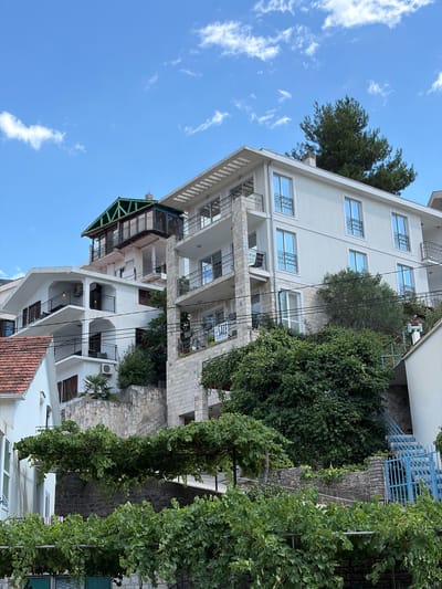 The image shows a multi-story residential building on a hillside in Lepetane, Montenegro. The building has white stucco walls and balconies with black railings. Several windows with blue-tinted glass are visible. The building is surrounded by lush green vegetation, including trees and what appear to be grapevines in the foreground. Stone walls and stairs are integrated into the landscape, indicating a terraced construction. A blue metal gate and stairs are visible on the right side of the image, leading up the hillside. The sky is blue with scattered white clouds. A "SALE" sign is attached to one of the balconies. There are no people visible in the image. The overall scene depicts a residential area in a Mediterranean coastal town.