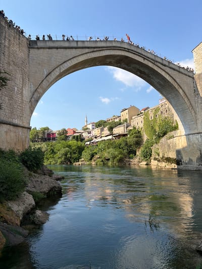 The image depicts a view from the Neretva River looking towards the iconic Stari Most (Old Bridge) in Mostar, Bosnia and Herzegovina. The bridge, a UNESCO World Heritage site, is constructed of stone with a large arch spanning the river. On top of the bridge, a crowd of spectators is gathered, observing an event. One individual, dressed in red shorts, is positioned at the edge of the bridge, appearing to be preparing to jump. The river flows calmly below, reflecting the blue sky and surrounding buildings. The riverbanks are lined with lush green vegetation and rocky outcrops. On the far side of the river, the historic old town of Mostar is visible, with its characteristic Ottoman-era houses painted in various colors, nestled on the hillside. A minaret of a mosque can be seen rising above the rooftops, indicating the city's rich cultural heritage. The scene is bathed in the warm light of a sunny day.