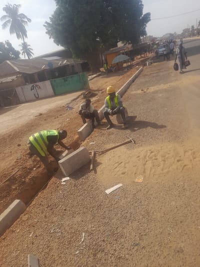 The scene depicts roadside construction in Ube, Nigeria, under bright daylight. In the foreground and midground, three adult males are present in an active construction zone. One man, wearing a lime-green high-visibility vest, dark shirt, and trousers, is positioned within an excavated dirt trench, actively placing or adjusting a grey concrete kerb stone. Adjacent to him, another man in a light-colored t-shirt and dark trousers is squatting near partially installed kerb stones. A third man, wearing a yellow hard hat, a lime-green high-visibility vest over a light shirt, and dark trousers, is seated on a concrete block or the ground further along the kerb line. A pickaxe with a wooden handle rests on the gravel-covered ground near the seated man.
The ground consists of reddish-brown dirt, excavated earth, gravel, and sand. A string line is visible, guiding the placement of the kerb stones. In the background, a paved road extends to the right, populated by numerous pedestrians, some carrying items or shaded by umbrellas. A woman in a light-colored outfit and carrying a dark bag walks prominently on the right side of the paved road. On the left, behind the construction area, stand several single-story buildings with corrugated metal roofs. One building features a white wall with a distinct red, white, and green "PDP" umbrella logo painted on it, adjacent to a green wall section. Multiple satellite dishes are visible on the roofs of these structures. Tall palm trees and a large, dense leafy tree are present in the background against a bright, possibly hazy sky.