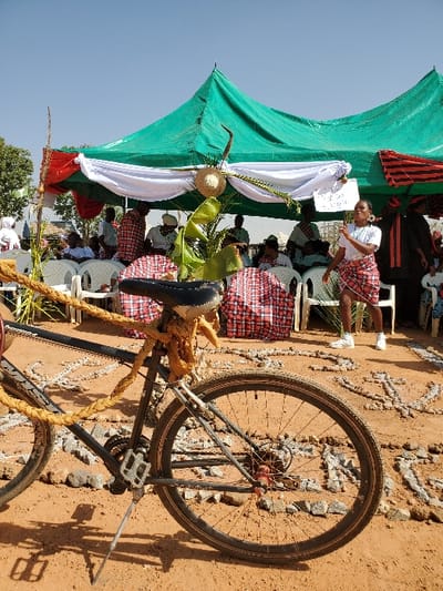 The scene depicts an outdoor event in Bar Gada, Nigeria. A black mountain bicycle with "GIANT" branding stands in the foreground on reddish-brown dirt, propped by a kickstand. The bicycle is decorated with a thick, braided tan rope wrapped around its frame and handlebars. Attached to the front of the bicycle are a bundle of green leaves, a small woven straw hat, and a curved, horn-like object.
In the midground, a large tent with a green tarp roof provides shade. The tent's front is decorated with white and red fabric drapes, along with green fronds and a small woven straw hat. Beneath the tent, numerous individuals are seated on white plastic chairs; some chairs have red and white checkered fabric covers. A young woman, wearing a white t-shirt, a red and white checkered skirt, and white sneakers, is standing in front of the tent, mid-movement, holding a white sign in her left hand. Other attendees in various attire are visible seated under the tent.
The dirt ground around the bicycle and extending into the midground is adorned with patterns, including partial letters and an anchor-like symbol, formed from light-colored stones or debris. The background consists of a clear blue sky and sparse trees on the left. The scene is illuminated by bright daylight.