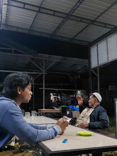 A low-angle shot shows two men at a table. One man, wearing a white skullcap, a black jacket, and a patterned scarf, gestures with his hands. The other man, in a blue sweatshirt, holds a smartphone and appears to be filming or photographing. A green plate sits on the table near a lighter. The setting appears to be an outdoor or semi-outdoor space with a corrugated metal roof. It is nighttime, and the area is illuminated by artificial lighting.