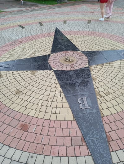 This is a ground-level, eye-level, medium shot of a compass rose pattern on a paved area in Goryachiy Klyuch, Russia. The compass rose is made of dark grey and black metal with inlaid letters and symbols. The paving stones are in concentric semicircles of pink, yellow, and light grey. Two people are visible in the upper right and upper left corners, walking on the paved area. The person on the right is wearing a red skirt and white shoes, and the person on the left is wearing dark pants and white shoes. The metal compass has a circular inset with Cyrillic text that reads "ГОРЯЧИЙ КЛЮЧ" (Goryachiy Klyuch). Another Cyrillic letter "Б" is visible on one of the arms of the compass. The ground appears wet, suggesting recent rain.
