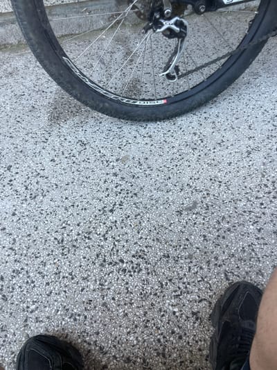 The image shows a close-up of a bicycle wheel and a person's feet on a speckled concrete surface. The bicycle wheel has a black tire with a white logo that reads "DISC". The rear derailleur is visible, as well as spokes and a chain. The concrete ground is composed of small, dark and light-colored pebbles embedded in a lighter matrix. The person is wearing black sneakers. The image appears to have been taken outdoors, possibly in an urban setting. There is no visual information to confirm the location as Haskovo, Bulgaria.