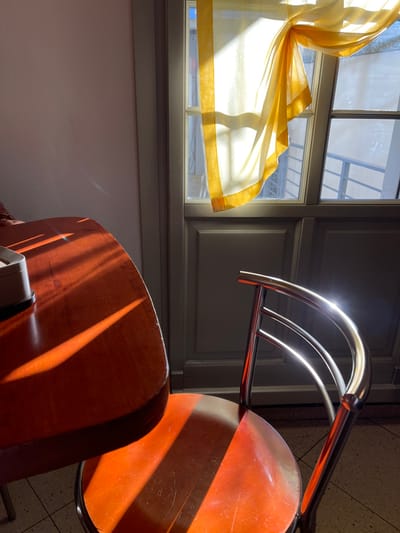 The image presents an indoor scene, likely a room in Sondrio, Italy, characterized by strong, warm sunlight, indicating either early morning or late afternoon. On the left, a multi-paned window is partially draped with a translucent, billowy yellow curtain, which is brightly illuminated and appears to be gently uplifted by a breeze or the angle of the light. Through the clear windowpanes, a soft view of exterior buildings or structures is visible.
To the right and bottom, elements of two chairs are prominently featured. These chairs possess wooden seats and backs, with sleek, curved chrome or metallic frames that gleam brightly where the sun hits them, creating noticeable highlights. Intense, elongated shadows are cast across the wooden surfaces of the chairs and the adjacent floor, accentuating the directional light. The window frame itself is a darker, muted tone, possibly gray or dark green, contrasting with the lighter, neutral-colored wall below it.
The scene is devoid of people or overt activity, creating a quiet and contemplative atmosphere. The vivid interplay of light and shadow, combined with the warm hues of the wood and the curtain, evokes a sense of tranquility and warmth. No visible text is present in the image.