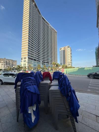 The image captures an outdoor scene in South Quy Nhon Ward, Vietnam, under a clear blue sky. In the foreground, stacks of grey plastic chairs are draped with blue fabric, some of which are tied with white bows. A red fabric is draped over one of the chairs. Behind the chairs, a white car is parked.
The middle ground features a street with parked cars and a pedestrian crossing. Further back, several modern buildings are visible, including a tall, multi-story building with a light-colored facade and numerous windows, and a shorter, lighter-colored building to its right. Palm trees are scattered throughout the landscape. A green construction barrier runs along the street.
In the distance, the sky is mostly clear with a hint of cloud. The overall impression is one of an urban environment, potentially preparing for an event or reflecting a recent one, given the draped chairs.