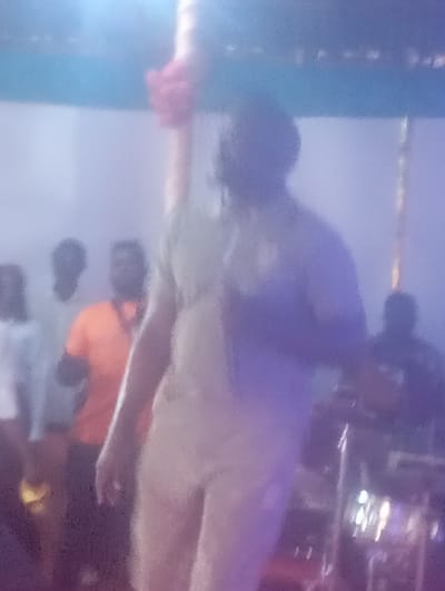A blurry image shows a man speaking into a microphone on a stage. The stage has several people on it, including one in an orange shirt. There appears to be a drum set on the stage to the right. The image seems to be taken at night, with stage lights illuminating the scene. The city of Markurdi, Nigeria, is not discernible.