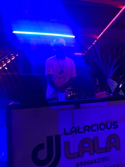 The image depicts a dimly lit indoor scene, likely a nightclub or event venue, bathed in vibrant blue and pink lighting. A person wearing a white cap and a light-colored t-shirt stands behind a DJ booth. The DJ booth has a large, stylized logo that reads "DJ LALA" in bold, outlined letters, with "LALACIOUS" printed above it. Below the logo, a phone number, "09066822115," is visible. The overall ambiance suggests a lively, possibly music-focused event taking place. The mention of Shendam, Nigeria, indicates the geographical location of this scene.