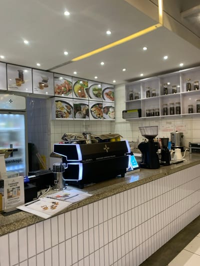 The image shows a counter in a cafe, located at coordinates -7.941 latitude, 112.608 longitude, which is in the city of Malang, Indonesia. The counter is made of light grey tiles and has a black and white espresso machine with the words "ALLEGRA MACCHINE ESPRESSO ITALIANE" on its front. There are several glass jars with coffee beans on a shelf above the counter. There are also several empty glass jars and a large black coffee grinder on the counter next to the espresso machine. To the left of the espresso machine are two large black cash registers and a sign that says "CASHLESS NOTICE". Behind the counter, on the wall, are multiple white framed pictures showcasing various types of food and coffee. The cafe has a white tiled ceiling with recessed lighting.