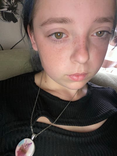 The image is a close-up, indoor selfie of a young person with light brown eyes and freckles. They have dark hair with streaks of blue. They are wearing a black, ribbed shirt with a cutout in the front and a silver chain with a pendant featuring a picture of a cat. The background includes a couch and a wall with a floral pattern. The lighting suggests an indoor environment. The location is not identifiable from the image.