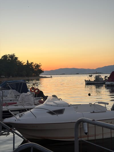 The image depicts a tranquil waterfront scene at sunset in Keramoti, Greece. The sky transitions from a pale blue at the top to a vibrant orange and yellow near the horizon, casting a warm glow on the water. Several boats are docked in the foreground and middle ground. In the immediate foreground, a white motorboat is prominently featured, with red lettering on its hull reading "ΔΕΣΠΟΙΝΑ Τ.ΚΕΡ.29". Another white boat with a blue canopy is docked behind it, and further back, a larger white boat with a cabin and a red canopy is visible. In the distance, across the calm water, a tree-lined shore is visible with mountains in the background. Two figures are standing on a pier in the middle distance, appearing to interact with the water or the boats. The overall atmosphere is peaceful and serene, characteristic of a Mediterranean sunset.