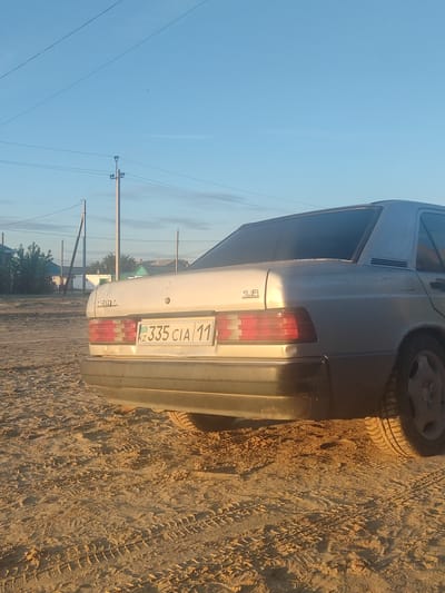 A 1980s Mercedes-Benz W201 sedan, possibly a 190E 1.8, is parked on a sandy, unpaved road in the city of Aral, Kazakhstan. The car is silver and appears dusty. The license plate reads "KZ 335 CIA 11". In the background, several houses and utility poles are visible. The sky is clear and blue, with the sun low in the sky, casting long shadows. The ground is covered in tire tracks.