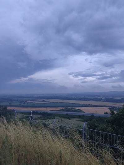 The image depicts a wide, panoramic view of a rural landscape under a dramatic, cloudy sky. In the foreground, dry, tall grasses and a metal fence with barbed wire are visible. Beyond the fence, a road or highway runs horizontally across the scene, bordered by fields of golden crops, dark green trees, and patches of lighter green vegetation. In the distance, the landscape softens into a hazy, undulating terrain with faint indications of human settlement or infrastructure, suggesting a vast agricultural area. The sky is filled with heavy, dark grey and blue clouds, hinting at an impending storm or the aftermath of one. The overall atmosphere is somber and natural, with a strong sense of open space and the power of weather. This scene could be situated in the vicinity of Svilengrad, Bulgaria, a region known for its agricultural lands and rolling hills, where such expansive views under changing skies are common. There are no people or animals visible in the image.