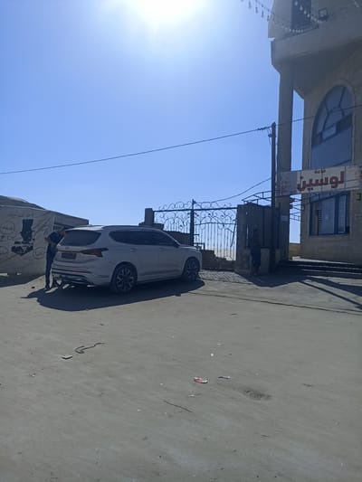 A white SUV is parked on a dusty, paved area in front of a metal gate. A man in a dark shirt and jeans stands next to the rear of the SUV, facing it. Behind the gate, a building with a cream-colored facade and arched windows is visible. A sign above an entrance to the building displays Arabic text that reads "قاعة لوسين" (Hall Lousin). The scene is bathed in bright sunlight, suggesting it is daytime. The sky is clear and blue. The ground is covered in loose dirt and debris. In the background, there are some tents or tarps. The overall atmosphere appears mundane, with no strong emotional cues evident. The time of day is likely late morning or early afternoon due to the strong sunlight and shadows. The location appears to be a commercial area or a venue.