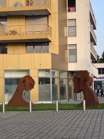 The scene depicts two rust-colored, abstract metal sculptures of human heads positioned on a strip of green grass, facing each other. These sculptures are situated in front of a modern building complex in Bischheim, France.

The building on the left features a golden-yellow facade with multiple stories. The upper floors have balconies with decorative, laser-cut metal railings depicting a naturalistic, branching pattern. The ground floor consists of large glass panels, some partially frosted, reflecting the surroundings. A white decal on one window displays a phone number "06 24 16 67 12" and a blue butterfly logo.

Adjacent to this, on the right, is a building with a white facade. This structure also has multiple stories with vertical rows of windows, some equipped with external blinds. On an upper floor, an open window reveals a folded pink towel hanging over the sill. Further to the right, a sign partially visible displays "Crédit," indicating a financial institution.

The foreground consists of a paved area made of rectangular pavers in a grid pattern. Four silver metallic bollards are evenly spaced on the edge of the paved area, separating it from the grass strip where the sculptures stand. In the mid-ground, beyond the "Crédit" sign, two indistinct human figures are visible walking from right to left. Sparse trees are present in the distant background on the right. The sky is clear, suggesting daytime conditions.