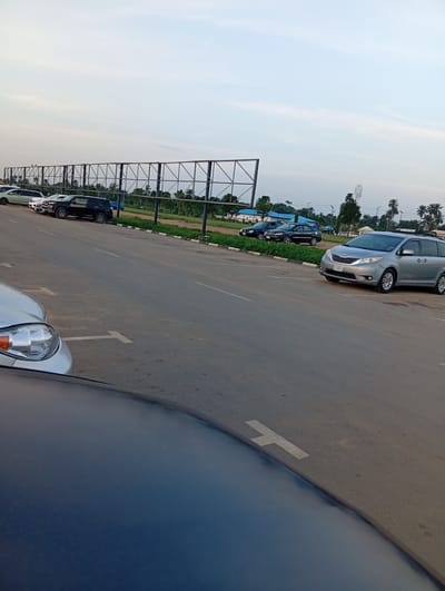 The image depicts a parking area in Omagwa, Nigeria. In the foreground, part of a dark-colored car's hood is visible. Further back, a silver minivan is parked along a road with painted parking markers. Other vehicles are parked in a row further down the road. A large metal structure stands along the road, possibly for billboards or signage. Trees and vegetation line the background. The sky is overcast.