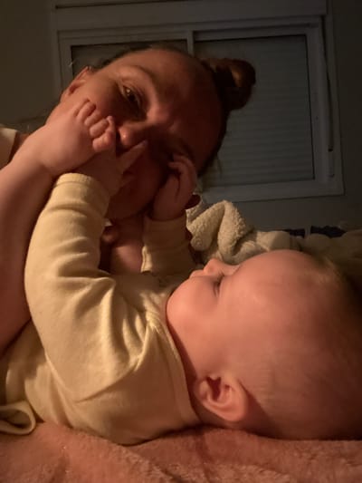 A woman and a baby are pictured indoors. The woman has her hair in a bun and is looking at the camera. The baby is lying on her chest and has its hand on her face, near her eye. The baby is wearing a beige long-sleeved shirt. A window is visible in the background on the left side of the frame. A blanket or cloth is partially visible behind the baby. The location of Florianópolis, Brazil, is not discernible from the image.