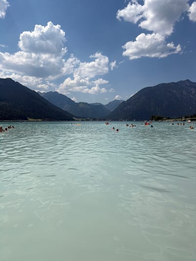 This media file depicts a scenic summer day at Lärchenwiese, Austria. The image is oriented vertically, with the sky and mountains on the left and a body of water on the right. The sky is a vibrant blue, dotted with fluffy white clouds, suggesting fair weather.

The primary subjects are the people enjoying the clear, pale blue water of a lake. There are numerous individuals visible, some swimming and others wading closer to the shore. The water appears calm with gentle ripples reflecting the sunlight. Along the shoreline, there is a stretch of green land dotted with trees and some buildings, possibly indicating a recreational area or village. In the background, imposing, dark green mountains rise dramatically, their slopes covered in dense forest. The sun appears to be high in the sky, casting bright light and creating a warm, inviting atmosphere.

Further out in the lake, a yellow paraglider is visible, adding a touch of activity and adventure to the scene. The overall impression is one of leisure and enjoyment, with people taking advantage of a beautiful day to cool off in the lake and admire the stunning natural landscape. The combination of the clear sky, pristine water, and majestic mountains creates a picturesque and serene setting. No visible text is present in the image.