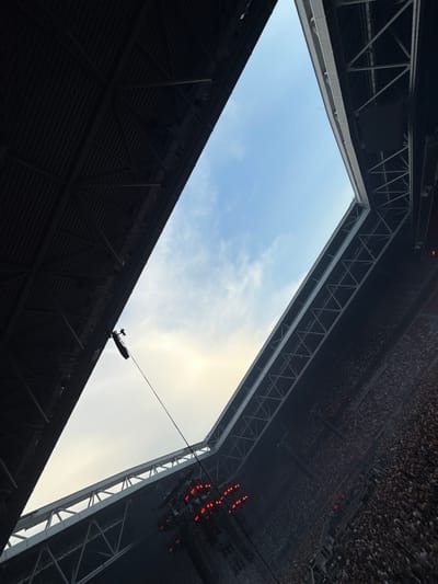This image was taken from the Esprit Arena in Dusseldorf, Germany, looking upwards at the sky through the open roof. A large crowd of people can be seen in the stadium seats, with a stage setup at the bottom right of the frame. Lighting rigs with red lights are visible above the stage. A cable is suspended from the stadium structure, with a black object attached to it. The sky is partly cloudy with a warm glow of sunset or sunrise.