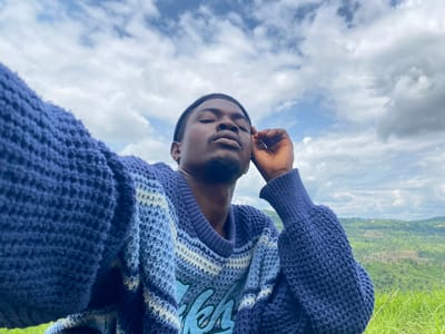 The image is an upside-down, close-up selfie of a young Black man wearing a blue knitted sweater with white and lighter blue stripes. He has short hair and a goatee, and his eyes are partially closed with a relaxed expression. His left hand is near his face, with his fingers gently touching his cheek. The background is a blurred landscape of green hills and a cloudy sky. The lighting suggests it is daytime. The image appears to have been taken outdoors in Achara, Nigeria, as indicated by the lush greenery and the open sky. There is no visible text in the image.