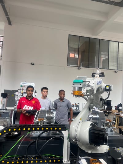 In a laboratory setting, three young men stand in front of a robotic arm. The man on the left, wearing a red Manchester United jersey with "AON" printed on it, holds a black controller. The middle man wears a grey t-shirt, and the man on the right wears a grey polo shirt. The robotic arm, labeled "SHSUNG", is white and has several attachments and sensors. In the background, there are other pieces of equipment, including a server rack with blinking lights, and large windows with blinds. The space appears to be a modern educational or research facility, likely located in Shuangfu, China, given the presence of Chinese text on some equipment labels, such as "SIMSUN" which is a Chinese robotics company.