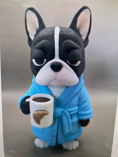 This is a close-up, vertically oriented, studio photograph of a small, decorative figurine of a Boston Terrier dog wearing a blue robe. The figurine is lying down, with its head turned slightly to the viewer's left. It has a black and white face with large, expressive eyes and a black nose. A white stripe runs down its muzzle.

The dog is holding a miniature white mug filled with a dark liquid, presumably coffee, and what appears to be a pastry or cookie with a croissant-like shape. The figurine is dressed in a plush, bright blue robe, cinched at the waist with a belt, and its paws are visible. The background is a neutral, textured surface, possibly a tabletop or a studio backdrop. The lighting appears to be soft and even, suggesting an indoor setting, likely for a product photo or a decorative display. There is no visible text in the image. The overall impression is of a whimsical and cozy scene.