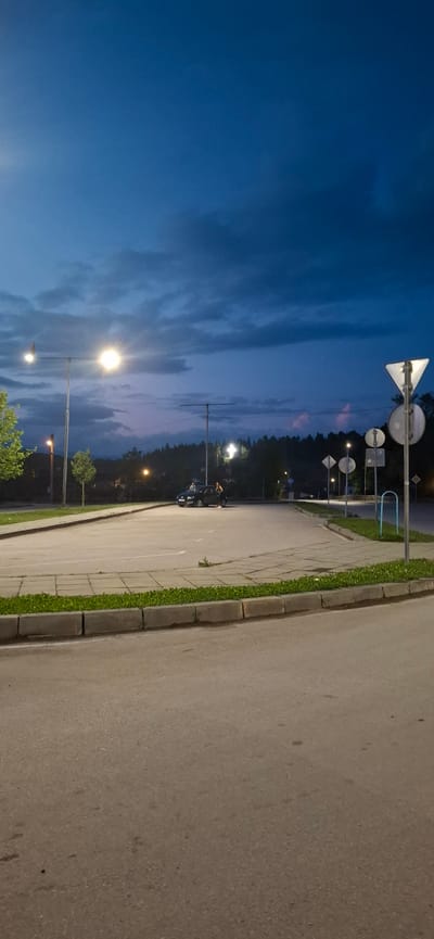 The image depicts an outdoor scene in the city of Bania, Bulgaria, at dusk or early night. The sky is a deep blue, transitioning from lighter blue at the upper left to darker, cloud-covered grey-blue across the majority of the frame, with faint pinkish-purple hues visible on the horizon line behind the distant treeline in the mid-right.
In the foreground, an asphalt road surface occupies the bottom quarter of the image. Adjacent to the road, on the right, is a concrete curb separating the asphalt from a strip of green grass, which in turn borders a paved area of rectangular concrete slabs. This paved area features faint white lines indicative of a parking lot.
Multiple artificial light sources illuminate the scene. A prominent, brightly glowing street light mounted on a tall pole is centered on the left side of the image. Another, less bright street light is visible further down the road on the left. Several additional street lights are discernible in the background, particularly along a winding road and within the distant forested area. Notably, a large, brightly illuminated cross-shaped structure is visible on the dark, forested hill in the background, slightly to the right of the image's vertical center.
A dark-colored sedan is parked on the concrete slab area in the mid-ground, facing towards the left. Two individuals are present near the car: one person is partially obscured near the front driver's side, and another person, wearing light-colored clothing on the lower half, is standing near the rear passenger side, appearing to interact with the vehicle.
Various road signs are visible on the right side of the image, mounted on poles. These include a large triangular yield sign, several circular signs, and rectangular signs. A blue, inverted U-shaped metal structure stands near the signs on the far right. Overhead wires or support structures extend horizontally from some of the light poles in the mid-ground. Green foliage, including a small tree on the far left and several smaller trees or bushes along the paved area, is present. A dense forest covers the hill in the background.