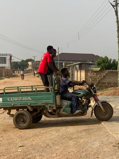 The image depicts a green three-wheeled motorized cargo vehicle, a "LUOJIA" brand, traversing an unpaved, dusty dirt road in Joma, Ghana. Four individuals are on board. A male driver, wearing a blue shirt and pants with white Crocs, is seated on the motorcycle portion, operating the handlebars. In the attached cargo bed, three male passengers are present. Two of them are standing upright; one wears a red t-shirt with a white '9' on the back and black shorts, while the other wears a dark t-shirt and dark shorts. A third passenger is partially visible, positioned lower in the cargo bed behind the other two.
The environment indicates a residential or semi-urban area. In the background to the left, a multi-story white building with dark windows is visible, along with other concrete block structures and utility poles supporting overhead wires. Further down the road, another motorcycle and individuals are discernible. To the right, a partially damaged concrete block wall stands alongside another utility pole. The sky is a light, hazy gray, suggesting an outdoor setting during daylight. The ground features dry, light-brown soil, with a visible concrete edge on the immediate right.