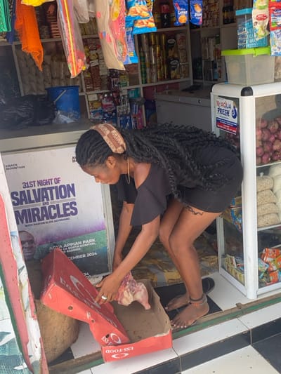 The scene depicts a woman interacting with a raw chicken in a red cardboard box at the entrance of a convenience store in Joma, Ghana. The woman, with dark skin and long black braids, wears a light-brown patterned headband, a black off-the-shoulder mini-dress, and is barefoot with a beaded anklet on her right ankle. A dark, swirling tattoo is visible on her left thigh. She is bent over, reaching into a red 'ESS FOOD' branded cardboard box containing a raw, plucked chicken. To her left, a large poster advertises 'GRACE MOUNTAIN 31ST NIGHT OF SALVATION AND MIRACLES WITH PASTOR ELVIS AYEMANG' scheduled for '31ST DECEMBER 2024' at 'AGYA APPIAH PARK'. The store interior, visible behind her, features shelves stocked with various packaged goods, including snacks, drinks, and toiletries. A white freezer and a clear display cooler holding red onions are also present, with a 'MC AQUA FRESH' label on the cooler. The floor at the entrance is tiled in a black and white checkerboard pattern.