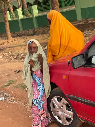 A red car is parked on a dirt lot. Next to it, a young girl is looking at the camera. She is wearing a colorful patterned dress and a light green head covering. Next to her is a woman in an orange covering. The car's tire is close to the girl, and the car's mirror is visible. The background shows trees and a green building. The scene appears to be outdoors during the day.