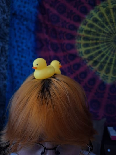 The image shows a close-up of a person's head, from the shoulders up, with two yellow rubber ducks perched on top of their head. The person has orange-tinted hair, and is wearing glasses. The background is a tapestry with a circular, mandala-like pattern in shades of blue, purple, and yellow. The setting does not provide any specific information to determine a location in Gyumri, Armenia.