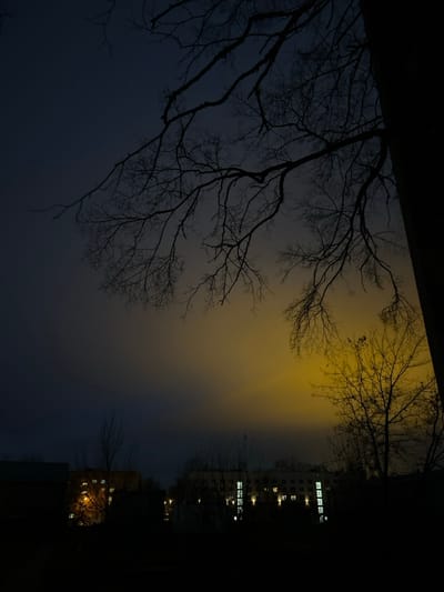 This nighttime image captures an urban or suburban landscape in Voskresensk, Russia. The sky transitions from a dark blue-grey in the upper left to a prominent, diffuse yellow-orange glow in the mid-right, suggesting significant light pollution or a powerful ground-level light source illuminating atmospheric haze or low clouds. In the foreground, numerous bare, deciduous tree branches are silhouetted against the sky, with a thick tree trunk visible vertically along the right edge. In the mid-ground, several multi-story buildings are present. A central, lighter-colored building exhibits multiple illuminated rectangular windows displaying white light. To its left, another building shows fewer windows with warmer, orange illumination. The immediate foreground is dark and indistinct, with faint lights visible at ground level between structures. No individuals or specific actions are discernible.