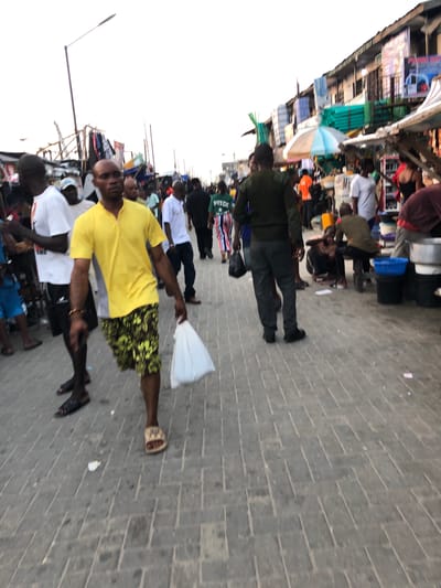 The image depicts a crowded street scene in Moba, Nigeria. The location appears to be a market area with stalls lining the left side of the frame. A variety of individuals are present, with most of them walking along the paved street. A man in a yellow shirt and floral shorts holds a white plastic bag. Street lights are also visible on the left.