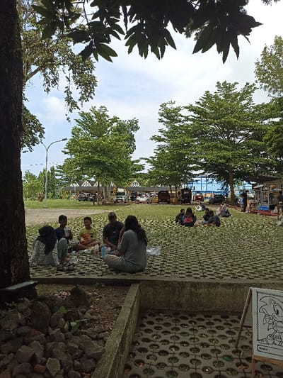 This outdoor scene captures a leisurely gathering in Mataram, Indonesia, likely in a park or recreational area. In the foreground, a group of about five to six individuals is seen sitting on a ground composed of paver blocks with grass growing through them. This group includes two adults – one man wearing a cap and another person with long dark hair seen from behind – and several children or young people, one in a yellow t-shirt and another in a striped top with a hijab. They appear to be picnicking or casually socializing, with plastic bags and wrappers suggesting food or drinks.
Further into the mid-ground and background, other smaller groups of people are also seated on the ground, indicating a public gathering spot. The area is well-shaded by numerous trees, with a prominent tree trunk on the left side of the frame. Beyond the grassy area, a parking lot is visible, filled with cars, and what appear to be small market stalls or vendor setups line the background.
The sky is bright with scattered clouds, typical of a sunny daytime, likely late morning or afternoon. The overall ambiance is relaxed and communal. In the bottom right corner, a small easel displays a coloring page featuring the character Sonic the Hedgehog, with the Indonesian word "NAMA:" (Name:) printed below, suggesting an activity for children.