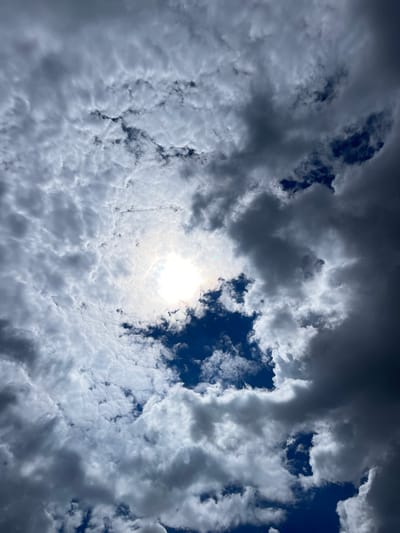 The image shows a sky filled with a mix of dark and light clouds. The sun is visible through a gap in the clouds, casting a bright light. The clouds appear textured and varied, with some areas being dense and dark, while others are lighter and more diffuse. The sky's color ranges from deep blue in the openings to shades of gray and white in the cloud formations. There are no people, subjects, objects, or actions depicted in this sky view.