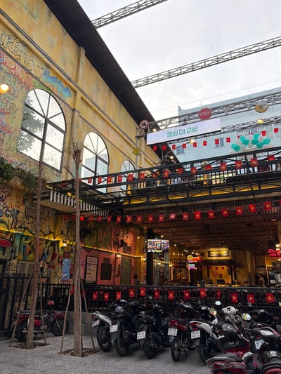 The image depicts a lively street scene in Thủ Đức City, Vietnam, featuring a building with vibrant graffiti on its exterior. The building has large arched windows and is adorned with rows of small Vietnamese flags. A canopy covers an outdoor seating area, where numerous motorcycles are parked on the sidewalk.
In the background, a large digital billboard displays text and images, with balloons in green and white suspended above. Signs with Vietnamese text are visible, along with a red stop sign. The sky is overcast, suggesting a cloudy day. Several thin trees with sparse foliage are planted in squares along the sidewalk.
The overall impression is one of a bustling urban environment, possibly a marketplace or a dining area, with a blend of traditional Vietnamese cultural elements and modern street art.