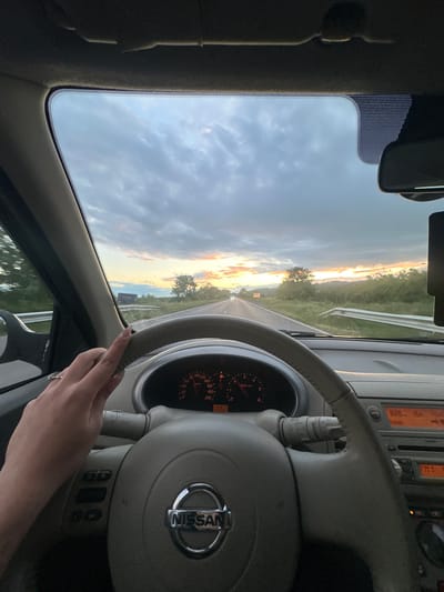 A view from inside a Nissan car, likely during a journey on a highway, perhaps near Padarevo, Bulgaria. The dashboard is visible, specifically the steering wheel with the Nissan logo. The speedometer displays the car's speed. A person's hand rests on the steering wheel, while the sunset illuminates the road ahead through the windshield. The sky is a mix of clouds and golden hues.