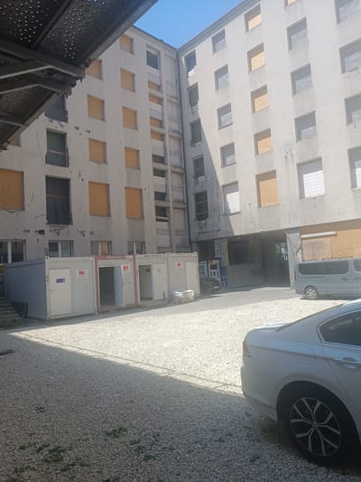 The image depicts a multi-story, dilapidated building with boarded-up windows, suggesting ongoing renovation or demolition. The facade is a light-colored concrete or stucco, with visible signs of wear and tear, including cracks and patches. Several windows are covered with plywood. Below the building, several prefabricated modular units or containers are lined up, appearing to serve as temporary site offices or storage. A light-colored sedan is parked in the foreground, partially visible on the right side of the frame, and a grey minivan is parked further back. The ground is covered with light-colored gravel. The scene is outdoors, under a clear blue sky, with shadows cast by the building and scaffolding. The setting appears to be an urban environment in Katowice, Poland, likely a construction or redevelopment site.