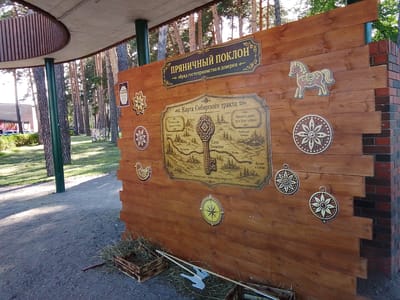 This image shows an outdoor informational display constructed from wooden planks. The display features a large, ornate sign at the top reading "ПРЯНИЧНЫЙ ПОКЛОН" (Gingerbread Bow), with a subtitle below it: "обряд гостеприимства и доверия" (rite of hospitality and trust).
Below the main sign is a detailed map titled "Карта Сибирского тракта" (Map of the Siberian Route). The map is illustrated with stylized drawings of settlements, churches, and landscapes, and depicts a winding path with key locations such as "Ялуторовск", "Село Уковское", and "Ирбитская ярмарка" (Irbit Fair). The map emphasizes the "Сибирский тракт - дорога добра и доверия" (Siberian Route - road of kindness and trust).
Various decorative elements are attached to the wooden wall, including stylized flowers, a small horse figurine, a chicken, and a compass rose. These elements likely relate to the theme of the display.
In the foreground, there are two wooden crates filled with straw or dried grass, and some long wooden poles, one of which appears to be part of a pitchfork. The ground is covered with gravel and dirt, and there is a pathway leading away. In the background, tall pine trees are visible, suggesting a park or forest setting. The lighting indicates it is daytime, likely late morning or afternoon, with sunlight filtering through the trees, casting shadows. The overall scene suggests an educational or tourist attraction in a rural or natural environment, possibly related to local traditions or history.