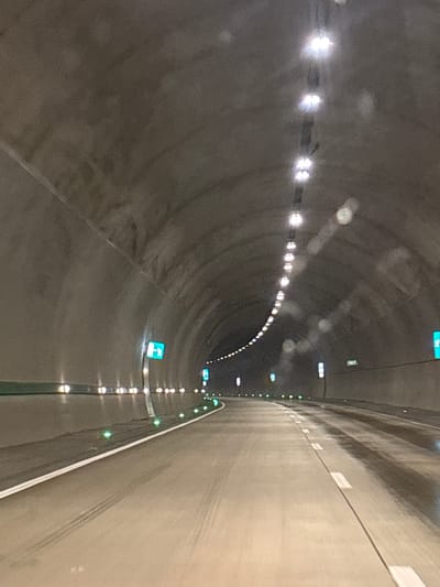 The image depicts the interior of a tunnel, likely a highway tunnel, given the lighting and road markings. The tunnel walls are constructed of rough concrete, and a series of lights are mounted along the ceiling, creating a illuminated pathway. The road surface appears to be asphalt and is wet, suggesting recent rain or maintenance. There are lane markings and smaller green lights embedded in the side of the tunnel, possibly for guidance or safety. In the distance, blue and white signs are visible, which are common in traffic infrastructure. The scene is devoid of people or vehicles, creating a sense of solitude. This tunnel is located in the vicinity of Ubisa, Georgia, a region known for its mountainous terrain and historical sites, which may influence the engineering and aesthetic of such infrastructure.
