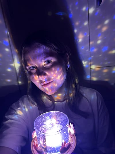An indoor scene in Gornyi (Dvurechenskoe S/P), Russia, depicts a young female in a dimly lit room. She is positioned centrally, gazing directly at the viewer with a slight smile. Her face, light-colored hair, and light-colored top are illuminated by numerous projected blue, white, and orange light patterns, resembling stars or circles. She holds a cylindrical light-projecting device with both hands at the bottom of the frame. This device emits the patterned light, and its top surface displays intricate white patterns resembling fireworks or snowflakes. Small, decorative elements resembling bunny ears are visible on the side of the device near her hands. The background consists of dark wall surfaces, also adorned with the projected light patterns and showing faint vertical seams.