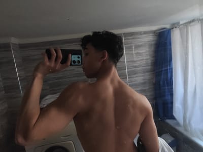 The image displays a young male capturing a mirror selfie within an indoor bathroom setting. The individual is shirtless, seen from the back, with his head turned to the right, looking over his shoulder. He possesses dark, curly hair with faded sides. A black smartphone, featuring multiple rear camera lenses, is held in his elevated left hand, angled to photograph his reflection. The immediate environment includes grey, horizontally striped wall tiles, a white shower curtain with a prominent blue section on the right, and a white washing machine partially visible in the lower-left background. A white towel is draped over an unseen object to the right of the individual's lower back. The specific location within As-Salihiya Municipality, Syria, is not ascertainable from the visual evidence presented.