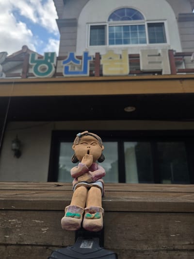 This is a medium shot, vertically oriented photograph taken outdoors in Gumi-si, South Korea. The image features a ceramic figurine of a child sitting on a wooden beam. The child is depicted with its eyes closed and hands near its mouth, suggesting a moment of thought or quiet. It is wearing a purple shirt and pink striped pants with purple and green accents on the boots.

In the background, to the left, is a building with a window and a circular decorative element above it. Large, stylized Korean characters are mounted on the building, some in blue and yellow. The characters appear to be part of a sign. The sky, with visible clouds, is reflected in the window. The wooden beam the figurine is sitting on appears weathered. The overall lighting suggests it is daytime, possibly late morning or early afternoon. The presence of the building with Korean text and the distinct architectural style points to a location in South Korea. No specific event is depicted, rather a still life composition of a figurine in its environment.