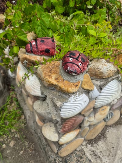 This image shows a close-up view of a decorative garden planter on a cloudy day, likely in Vladivostok, Russia. The planter is constructed from concrete and adorned with various seashells and smooth, rounded stones. Two small, painted rocks resembling ladybugs, with red bodies, black spots, and white eyes, are placed on top of the planter amongst green foliage. The planter itself is embedded with numerous seashells, predominantly white with ridged patterns, and some smaller, rounded stones. The surrounding environment consists of more greenery and a greyish, textured ground surface. The composition is focused on the planter's artistic details and its integration with the natural elements of the garden.