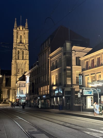 The image captures a nighttime street scene in Ghent, Belgium, focusing on the Sint-Niklaaskerk (Saint Nicholas' Church) tower and surrounding buildings. The church tower, illuminated by artificial lights, is a prominent feature on the left side of the frame, exhibiting Gothic architectural style with spires and arched windows. Adjacent to the tower, a series of buildings line the street, displaying various architectural elements and illuminated windows.
In the foreground, tram tracks run along the asphalt road, indicating active public transportation. Several streetlights and overhead wires for the tram system are visible, contributing to the urban nighttime ambiance.
On the sidewalk, several individuals are present. A few people are standing near a bus stop or information display, which features text and numbers, possibly indicating public transport schedules. One person appears to be a woman in a light blue dress and another is a man in a light-colored jacket and dark pants. Further down the street, a group of people are gathered near a building entrance, and some individuals are on bicycles.
Businesses with illuminated signs, such as "JOEY" and "Leonidas," are visible along the street, suggesting commercial activity. The overall atmosphere is one of a vibrant, well-lit European city at night, with a blend of historical architecture and modern urban life. The sky is dark, indicating late evening or night.