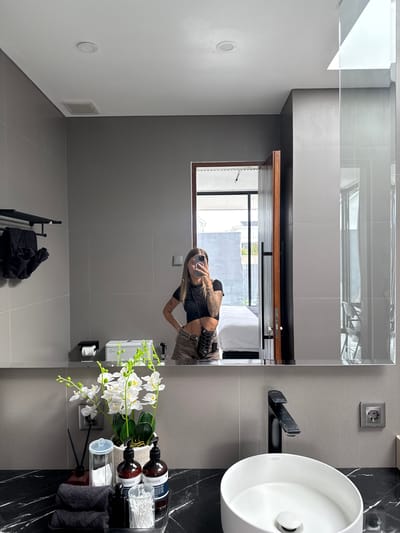 In a modern bathroom in Kediri, Indonesia, a person is taking a selfie in a large mirror. The bathroom features gray tiled walls, a white ceiling with a recessed light, and a black marble countertop. On the countertop, there's a white vessel sink with a black faucet, a white orchid plant, several bottles of soap and lotion, cotton swabs, and folded black towels. To the left, a black towel rack holds additional folded towels. In the reflection of the mirror, a doorway leads to another room with a bed and a window. The person in the selfie is wearing a black crop top and patterned shorts, with visible tattoos on their arms. An electrical outlet is visible on the tiled wall.