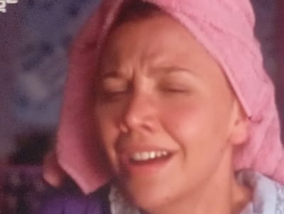 The image is a close-up, slightly blurry, and grainy photograph of a person's face from the chest up. The subject is wearing a pink towel wrapped around their head, covering their hair. Their eyes are closed or squinting, and their facial expression suggests discomfort or a grimace. The person's mouth is open, revealing teeth, and their skin appears flushed. The background is out of focus and shows indistinct dark and colored shapes, potentially suggesting an indoor environment. There is no discernible information in the image that would indicate the location of Sofiivska Borshchahivka, Ukraine.
