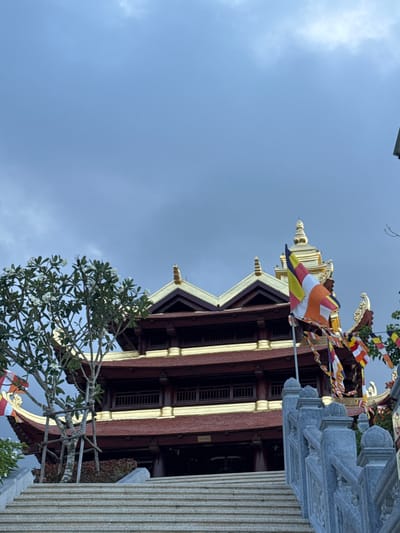 The image shows the exterior of a Buddhist temple, likely located in Diên Khánh, Vietnam. The temple features traditional architecture with tiered, dark brown wooden roofs trimmed in gold, and a prominent golden spire. Several colorful prayer flags are strung across the scene, fluttering in the wind. A stone staircase leads up to the temple entrance, flanked by ornate stone railings. To the left of the stairs, a flowering tree with white blossoms adds a touch of greenery. The sky is overcast with gray clouds, suggesting a muted or impending weather change. The scene is devoid of people, focusing entirely on the architectural and atmospheric elements of the location.