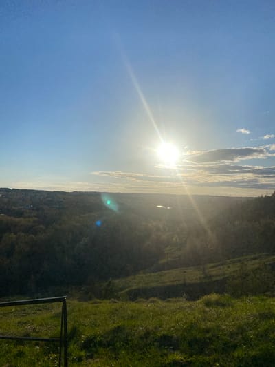 A landscape image taken outdoors during the daytime. The sky is predominantly light blue with wisps of clouds near the horizon. The sun is bright and creates lens flares. Below the horizon, there is a view of a hill or mountain covered in vegetation. The vegetation appears to be a mix of trees and grass. There is a dark metal fence in the bottom right corner of the image. The image is vertically oriented, with the sky at the left and the ground at the right. There are no people in the image. The location of the city Karakulino, Russia is not clear.
