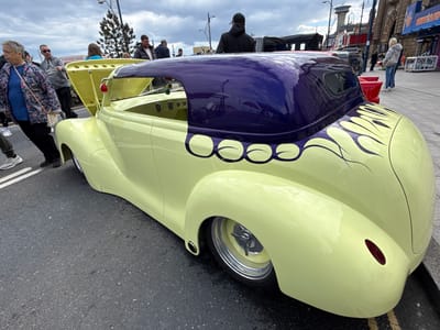 The image shows a custom-built, pale yellow hot rod with a dark purple roof and flame decals parked on a street in Great Yarmouth, United Kingdom. People are seen walking around and observing the car. The hood of the car is open. In the background, buildings, street lights, and signage suggest a seaside resort location. One sign reads "CASINO SLOTS." A woman with a floral jacket is standing close to the car on the left. Other onlookers stand or pass by on the pavement and road. The sky is overcast.